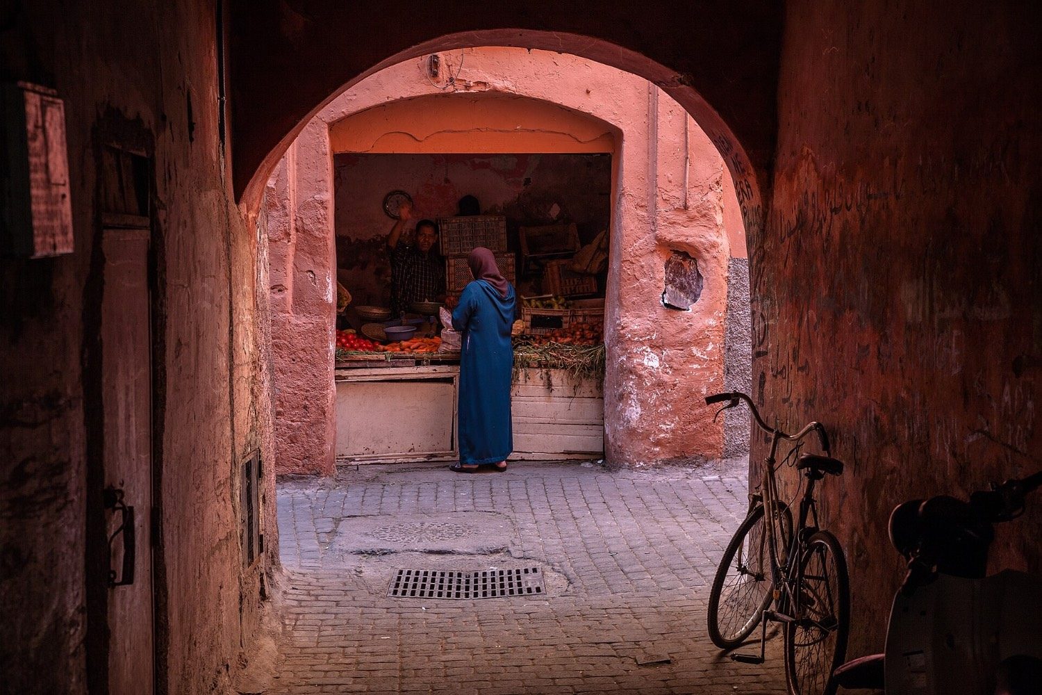 A woman in a long blue robe buys fruit and vegetables at a stall, seen through a pink-red archway in a narrow lane, with a bicycle leaning on the wall